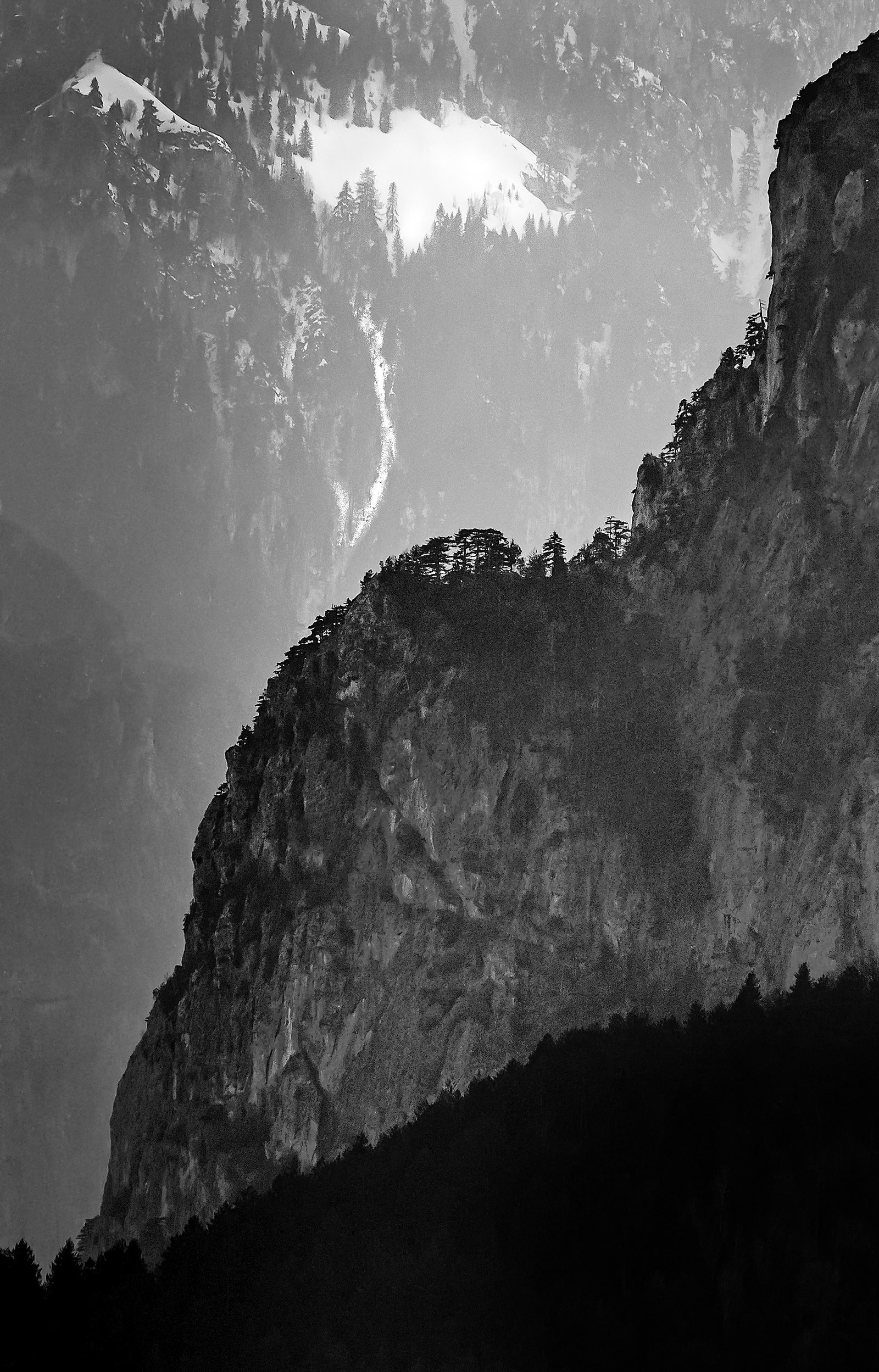 Vertical rock cliff rising above dark pine forest with snow-covered mountain slope and frozen waterfall in mist – limited edition fine art photography from the Existential Forms series by Aris Dimakopoulos